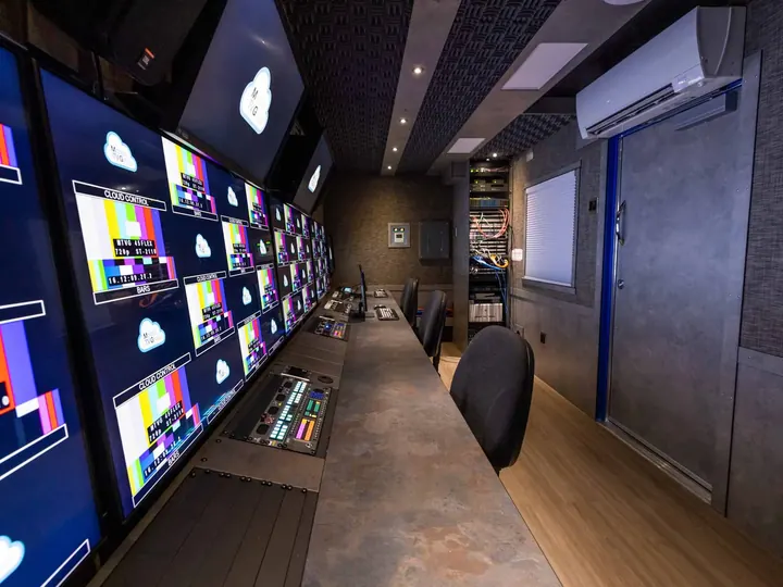 Container broadcast control room with wall of monitors, long console desk, mini-split AC and equipment rack
