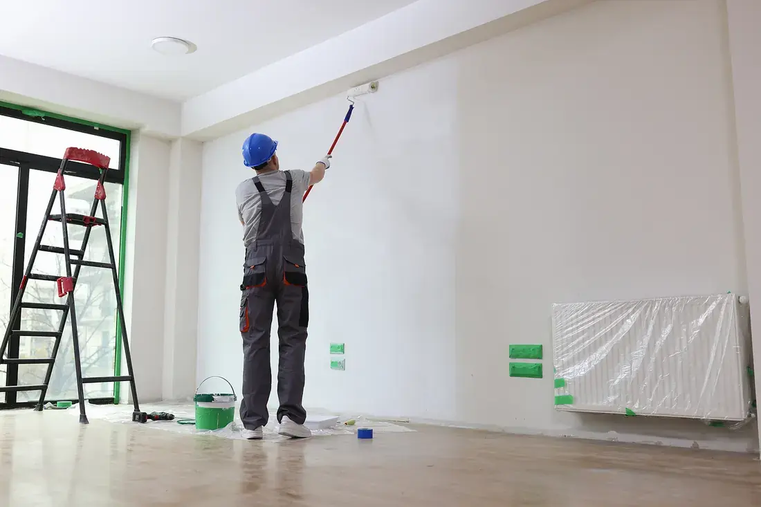 Painter rolling white paint onto a living room wall with the radiator masked off